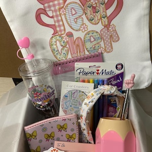 May include: A gift box filled with teacher-themed items. Includes a tumbler, notepad, pens, a pencil-shaped pen holder, a wallet, a headband, and a tote bag with the word "Teacher" printed on it in a floral design.