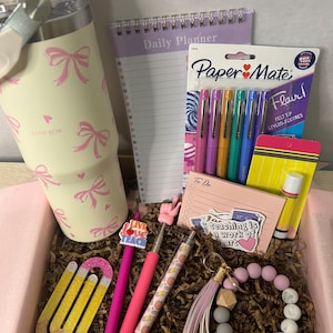 May include: A gift box filled with stationery and accessories. Includes a cream-colored tumbler with pink bows, pens, a notepad, Paper Mate Flair pens, a pencil-shaped paper clip, a bracelet, and a keychain. The notepad says "Daily Planner".