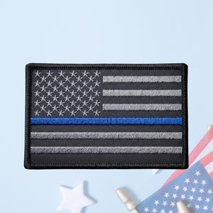 May include: A rectangular embroidered patch featuring a stylized American flag design. The flag is rendered in shades of black and grey, with a single horizontal blue stripe running across the center. The patch has a thick black stitched border.
