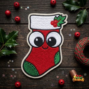 May include: A red and white embroidered Christmas stocking patch featuring large cartoon eyes, a white cuff, and a green toe section. The design includes a small sprig of holly with red berries on the cuff and a stitched smile on the stocking.