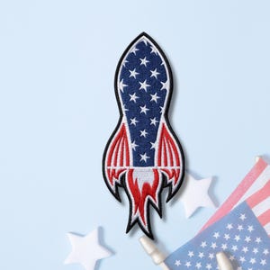 May include: An embroidered patch shaped like a rocket ship featuring a blue background with white stars on the main body, red and white vertical stripes on the fins, and a red and white flame design extending from the base with a black border.
