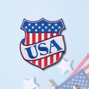 May include: An embroidered shield-shaped patch featuring a blue upper section with white stars, red and white vertical stripes below, and a white diagonal banner across the center with the blue text USA, outlined in red and blue thread.