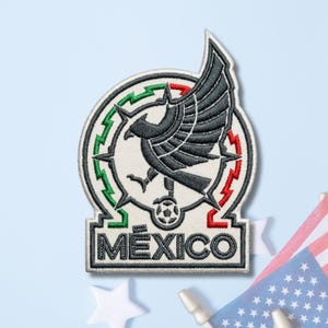 May include: An embroidered patch featuring a black eagle with spread wings perched on a soccer ball, centered within a circular frame outlined in green and red zig-zag patterns. The word MEXICO is stitched in black capital letters at the base.
