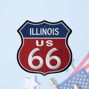 May include: A shield-shaped embroidered patch featuring a blue top section with the white text ILLINOIS and a red bottom section with the white text US 66. The patch has a black border and a small hole at the top center for hanging or attachment.
