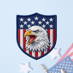 May include: A shield-shaped embroidered patch featuring a bald eagle head with a yellow beak and open mouth, set against a background of red and white vertical stripes and a blue upper section adorned with white stars, all bordered by dark blue thread.