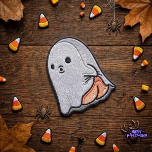 May include: An embroidered patch featuring a white ghost with black eyes and a small mouth, holding a piece of orange, yellow, and white candy corn. The patch is surrounded by scattered candy corn pieces, small plastic spiders, and dried leaves.