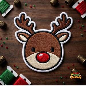 May include: An embroidered patch featuring the head of a reindeer with brown fur, large antlers, and a bright red nose. The design is outlined in white and black thread, showcasing a circular face shape with small black eyes and light tan accents.