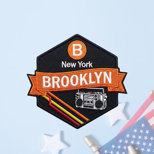 May include: A hexagonal embroidered patch with a black background featuring an orange circle with a white letter B, the text New York, and a banner reading BROOKLYN in white. Below the banner is a white boombox graphic and three diagonal stripes.