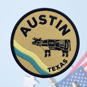 May include: A circular embroidered patch with a tan background and black border. The text AUSTIN appears at the top and TEXAS at the bottom in black. A black cow silhouette features labeled meat cuts, and a diagonal teal and yellow stripe.