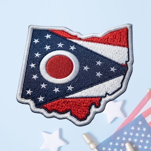 May include: An embroidered patch in the shape of the state of Ohio, featuring the Ohio state flag design. The patch displays a blue triangle with a white-bordered red circle and seventeen white stars, set against red and white horizontal stripes.