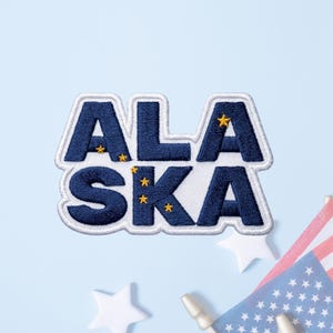 May include: A rectangular embroidered patch featuring the word ALASKA in bold dark blue block letters with a white border. Small yellow embroidered stars are integrated into the letters, mimicking the constellation design of the state flag.