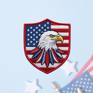 May include: A shield-shaped embroidered patch featuring a bald eagle head in profile with white, brown, and yellow details. The background of the patch displays the American flag with red, white, and blue stripes and white stars on a blue field.