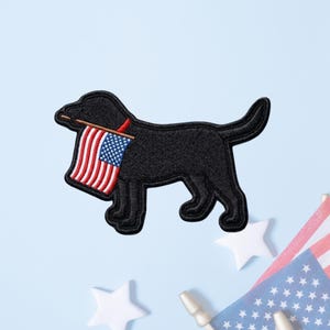 May include: A black embroidered dog patch featuring a side profile of a canine holding a small American flag in its mouth. The flag displays the traditional red, white, and blue colors with a blue field of white stars and vertical red and white stripes.