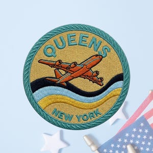 May include: A circular embroidered patch featuring a tan background with a teal border. The text QUEENS is arched at the top and NEW YORK at the bottom in teal. An orange airplane is centered above wavy black, light blue, and yellow lines.