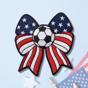 May include: An embroidered patch shaped like a bow with a soccer ball at the center. The top loops feature a dark blue background with white stars, while the bottom tails display red and white vertical stripes, all outlined in black thread.