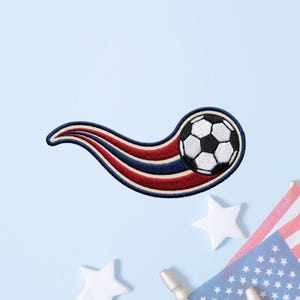 May include: An embroidered patch featuring a black and white soccer ball with a trailing motion effect in red, white, and navy blue stripes. The design is curved and outlined in navy blue thread, creating a dynamic appearance for this sports accessory.