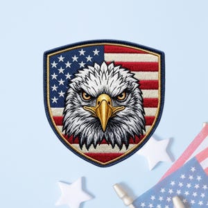 May include: A shield-shaped embroidered patch featuring a detailed bald eagle head with a yellow beak centered over a background of the American flag with red and white stripes and a blue field with white stars, all enclosed by a dark blue border.