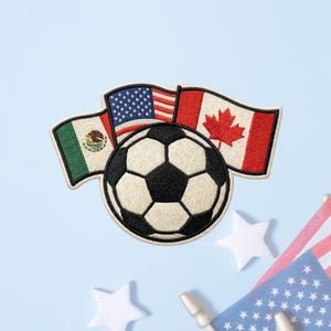 May include: An embroidered patch featuring a central black and white soccer ball positioned in front of three waving flags: the green, white, and red flag of Mexico, the red, white, and blue flag of the United States, and the red and white flag of Canada.