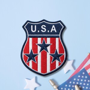 May include: A shield-shaped embroidered patch featuring a dark blue top section with the white text U.S.A. The lower portion displays vertical red and white stripes with three dark blue stars arranged in a triangular pattern across the center.