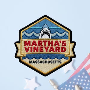 May include: A hexagonal embroidered patch featuring a shark emerging from blue ocean waves. A gold banner across the center displays the red text MARTHA'S VINEYARD, while the word MASSACHUSETTS is stitched in black at the bottom of the patch.