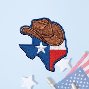 May include: An embroidered patch shaped like the state of Texas, featuring a brown cowboy hat resting on top. The patch displays the Texas flag design with a blue section containing a white star, a white vertical stripe, and a red vertical stripe.