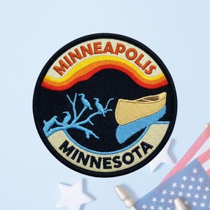 May include: A circular embroidered patch featuring a black background with the text MINNEAPOLIS at the top and MINNESOTA at the bottom. The design includes a yellow canoe on blue water, light blue birds on a branch, and orange and yellow stripes.