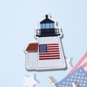 May include: An embroidered patch shaped like a lighthouse with a white tower, a dark blue lantern room, and a red roof on the attached building. The tower features a prominent American flag design with red and white stripes and a blue field with stars.