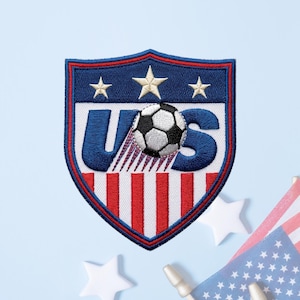 May include: A shield-shaped embroidered patch featuring the letters US in blue, with a black and white soccer ball replacing the letter O. The design includes three white stars at the top and red and white vertical stripes at the bottom.