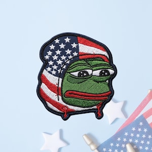 May include: An embroidered patch featuring a green frog character with large eyes and a red mouth, wearing a hood patterned with the American flag design of white stars on a blue field and red and white stripes, outlined in a dark navy blue border.