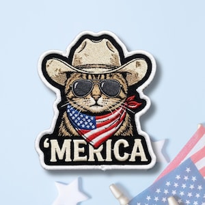May include: An embroidered patch featuring a brown tabby cat wearing a beige cowboy hat and black sunglasses. The cat has a red, white, and blue American flag bandana around its neck. The word 'MERICA is stitched in white at the bottom.