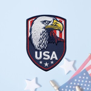 May include: A shield-shaped embroidered patch featuring a detailed white-headed eagle profile with a yellow beak. The background includes red and white vertical stripes, a dark blue base, the white text USA, and three white stars at the bottom.