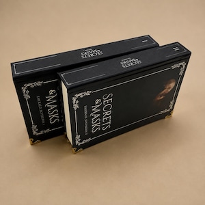 May include: Two black rectangular books titled Secrets & Masks Emerald Slytherin I and II. The covers feature white text, ornate white floral borders, and gold-toned metal corner protectors. The spines are also labeled with the book titles.