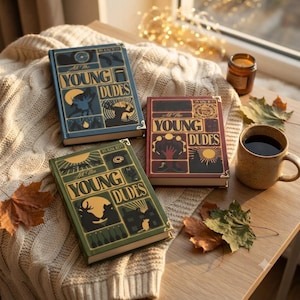 May include: Three hardcover books titled All the Young Dudes by MsKingBean89, featuring illustrated covers in blue, red, and green. Each book displays a grid of graphic panels with silhouettes of animals, objects, and celestial elements.