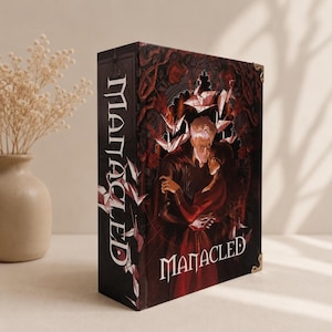 May include: A hardcover book titled Manacled with a dark, atmospheric cover illustration featuring two figures in an embrace surrounded by red roses and white origami cranes. The book spine and front cover display the title in a stylized white font.