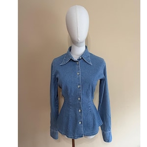 May include: A medium blue denim button-down shirt featuring a pointed collar, long sleeves with buttoned cuffs, and a fitted waistline that flares slightly at the hem. The shirt has a central vertical row of light-coloured buttons down the front.