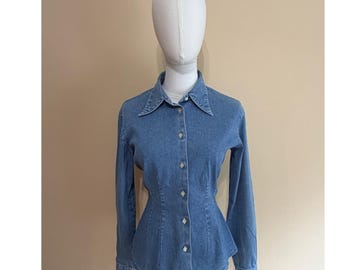 May include: A medium blue denim button-down shirt featuring a pointed collar, long sleeves with buttoned cuffs, and a fitted waistline that flares slightly at the hem. The shirt has a central vertical row of light-colored buttons down the front.