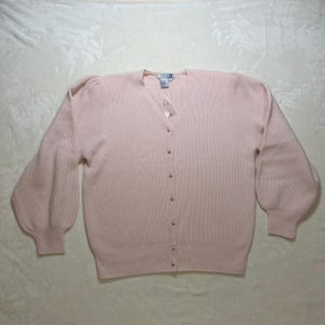 May include: A light pink, long-sleeved cardigan sweater featuring a V-neckline, ribbed knit texture, and a front button closure with six small, round, pearlescent buttons. The sleeves are designed with a slight puff at the shoulder and ribbed cuffs.