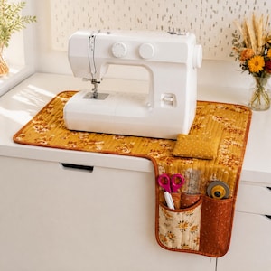 May include: A white sewing machine sits on a quilted mustard yellow mat with a floral pattern. The mat features an attached pocket organiser containing pink scissors, a white pen, and a rotary cutter, along with a matching square pincushion.
