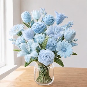 May include: A bouquet of light blue fabric flowers in a clear glass vase. The arrangement includes various blossoms such as roses, lilies, and daisies, all featuring soft blue petals and green fabric stems and leaves, creating a dense floral display.