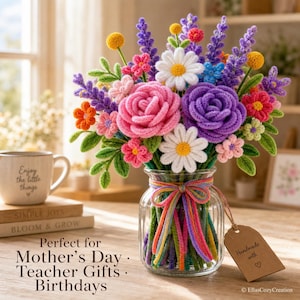 Könnte beinhalten: Ein Strauß aus gehäkelten Blumen in einem klaren Glasgefäß, das mit einem bunten Garnband gebunden ist. Das Arrangement enthält rosa und violette Rosen, weiße Gänseblümchen sowie kleine orange, gelbe und rosa Blüten mit grünen Blättern. Ein braunes Etikett trägt die Aufschrift Handmade with a heart.