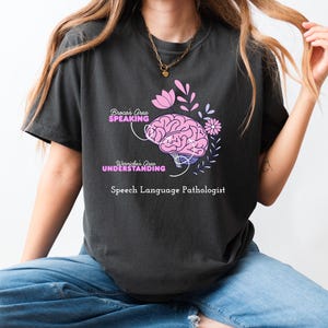 May include: Dark grey t-shirt with a pink brain graphic and the text "Broca's Area SPEAKING" and "Wernicke's Area UNDERSTANDING". The shirt also includes the words "Speech Language Pathologist".