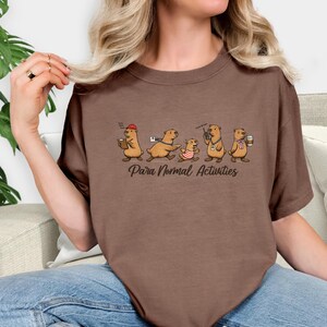 Puede incluir: Camiseta marrón con una ilustración de dibujos animados de tuza. Las tuzas se representan en diversas actividades, como sosteniendo tazas de café y usando un walkie-talkie. El texto "Para Normal Activities" está impreso debajo.
