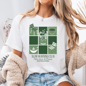May include: White t-shirt with a green grid design featuring coffee cups, chairs, and flowers. The text "SLOW MORNING CLUB" is printed below the grid, along with the phrases "Slow down, be Present" and "Drink Plenty of Coffee."