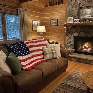 Könnte beinhalten: Ein gemütliches Wohnzimmer mit einem braunen Ledersofa, das mit einer grob gestrickten amerikanischen Flaggen-Decke drapiert ist. Kissen in verschiedenen Farben und Mustern sorgen für Komfort. Ein Steinkamin mit Feuer und ein Fenster mit Schneeblick vervollständigen die warme Atmosphäre.