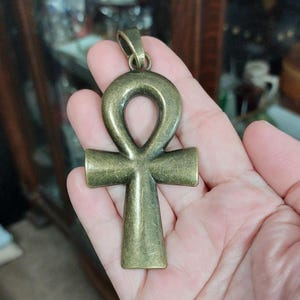 May include: A bronze-colored ankh pendant with a loop at the top and a cross shape below. The pendant has a vintage, antique appearance. The ankh is a symbol of life and immortality, often associated with ancient Egyptian culture.