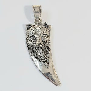 May include: Silver-toned pendant featuring a detailed wolf head design. The pendant is shaped like a stylized tooth, with intricate fur detailing and a bail for attaching to a chain. The pendant is made of a shiny, metallic material.