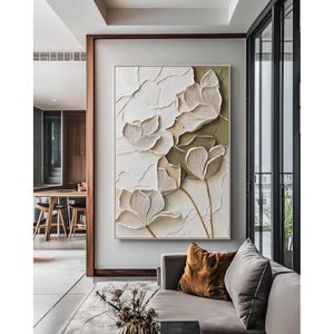 Pode incluir: Uma grande obra de arte de parede retangular com um design em relevo de flores estilizadas em tons de branco, creme e verde-oliva. A obra de arte é emoldurada em uma borda dourada e pendurada em uma parede cinza claro. Adequada para decoração.