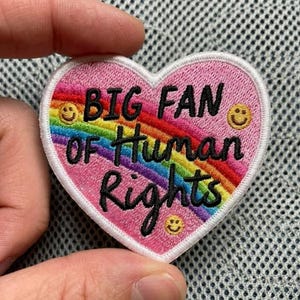 May include: Pink heart-shaped patch with the text "BIG FAN OF Human Rights" in black script. A rainbow arches across the center. The patch has three yellow smiley faces. The patch is white-bordered.
