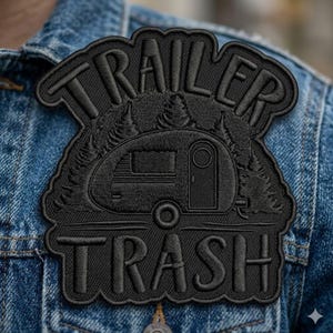 May include: Black embroidered patch with the words "TRAILER TRASH" above and below a camper illustration. The patch is in the shape of a camper and trees. The patch is sewn onto a denim jacket.