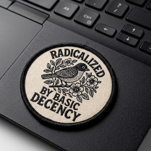 May include: A round patch with the text "RADICALIZED BY BASIC DECENCY" encircling a detailed black and white illustration of a bird perched on a floral branch. The patch is on a black laptop.